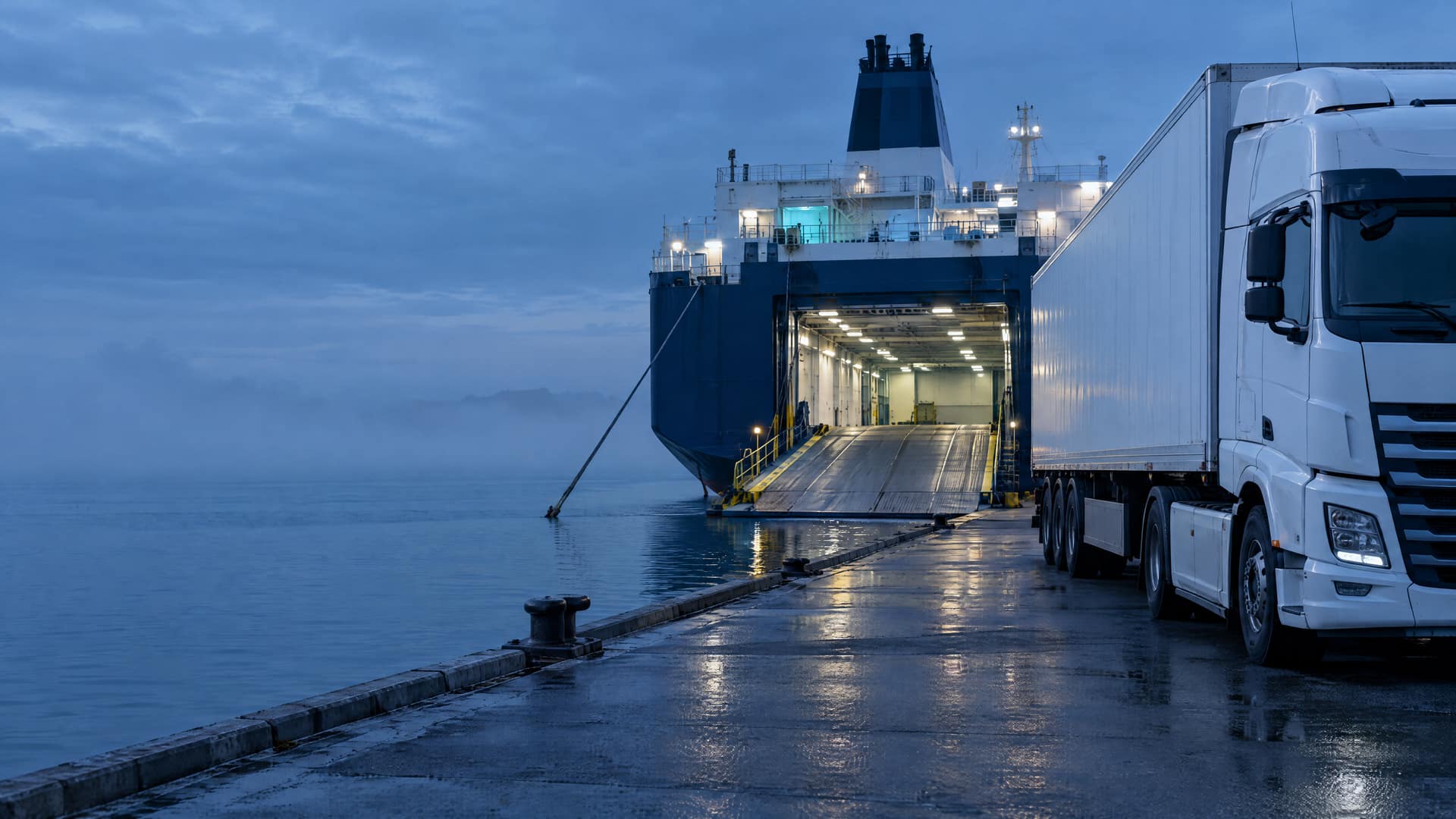 White freight truck on a quay beside a roll-on/roll-off ferry with its stern ramp lowered, at blue hour